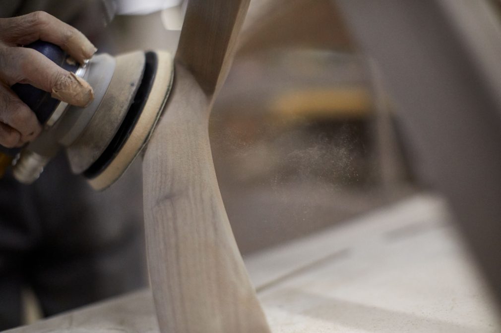 The image of a hand with a sanding tool. The tool is applied to the wood chair frame.