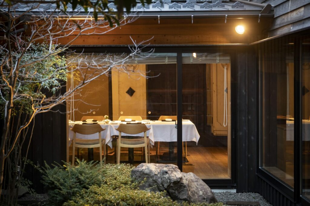 A dining room of a Japanese classic-style hote. A huge dining table with a white cloth is seen through a big window from outside. 