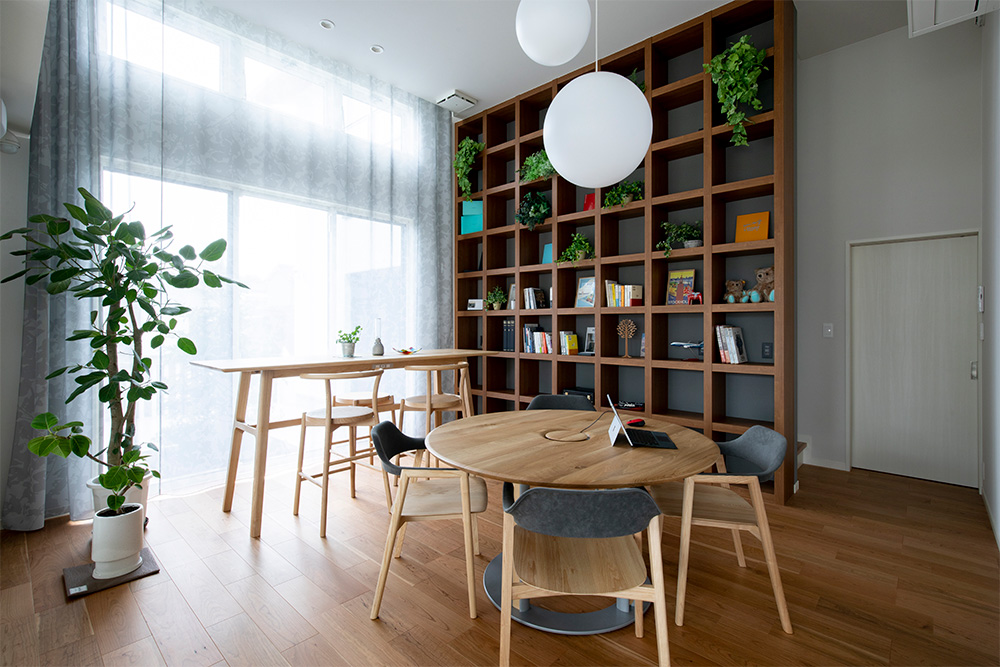 A room with a huge window. There's a high table with two dining chairs by the window, and a round table with four dining chairs.