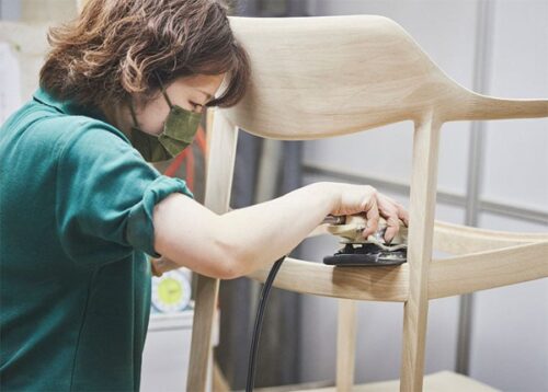 A woman is working on a wooden chair frame with an electric sander.