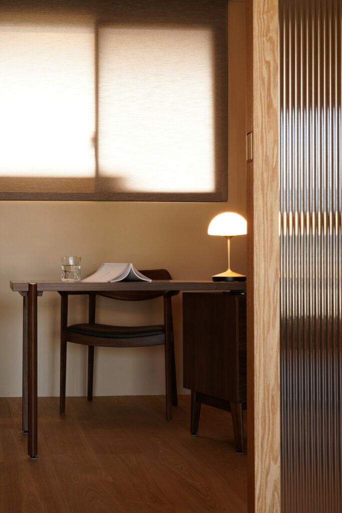 A wooden simple desk and a chair by the window. There's a simple lamp on the desk.