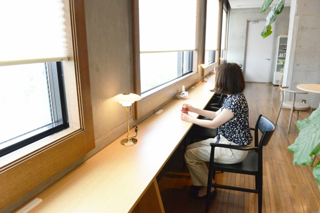 In a cafe area, there is a long table on the window side with a woman on a chair.