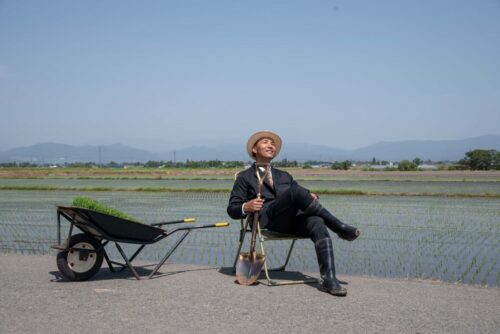 A guy in a business suit is sitting on a folding chair in the middle of rice fields.