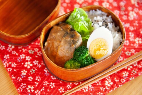 Box lunch, in which rice, chicken, a half-size boiled egg, and some vegetables are seen.