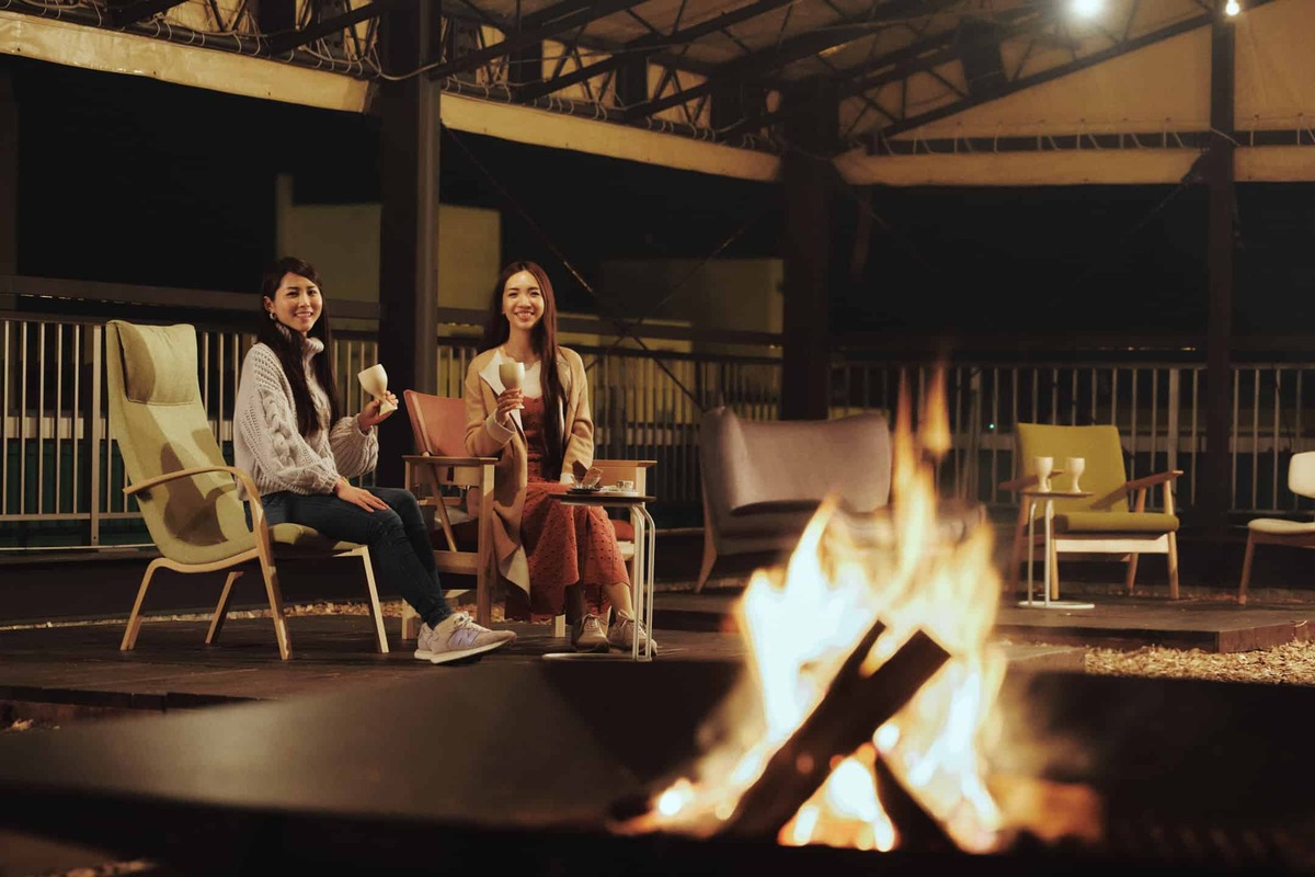 Two women are sitting on chairs and enjoying some drinks wine over bonfire.